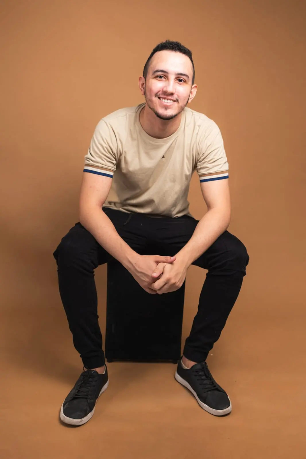 Homem jovem sorridente sentado em banquinho, usando camiseta bege e calça preta, com fundo marrom, expressão amigável e postura casual.