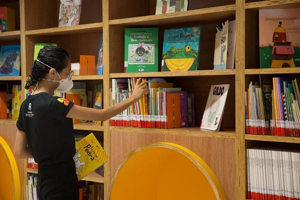 Na imagem, uma mulher, de máscara, em uma biblioteca, está com um livro amarelo nas mãos enquanto se estica para colocar outro livro em uma prateleira, que está cheia de livros infantis. O espaço tem estantes de madeira claras e assentos curvos e amarelos. No canto superior esquerdo da imagem, há um carimbo d'água com o texto 