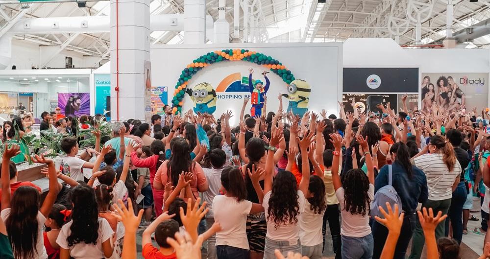 Na imagem, um grupo grande de crianças e adultos está reunido em um shopping center, todos com os braços levantados empolgados. No fundo, há um palco decorado com balões (nas cores verde, laranja e azul) e com personagens infláveis dos Minions. Uma pessoa fantasiada (talvez um palhaço ou animador) está no centro do palco, gesticulando para a multidão. O evento parece ser uma celebração ou show com o tema 