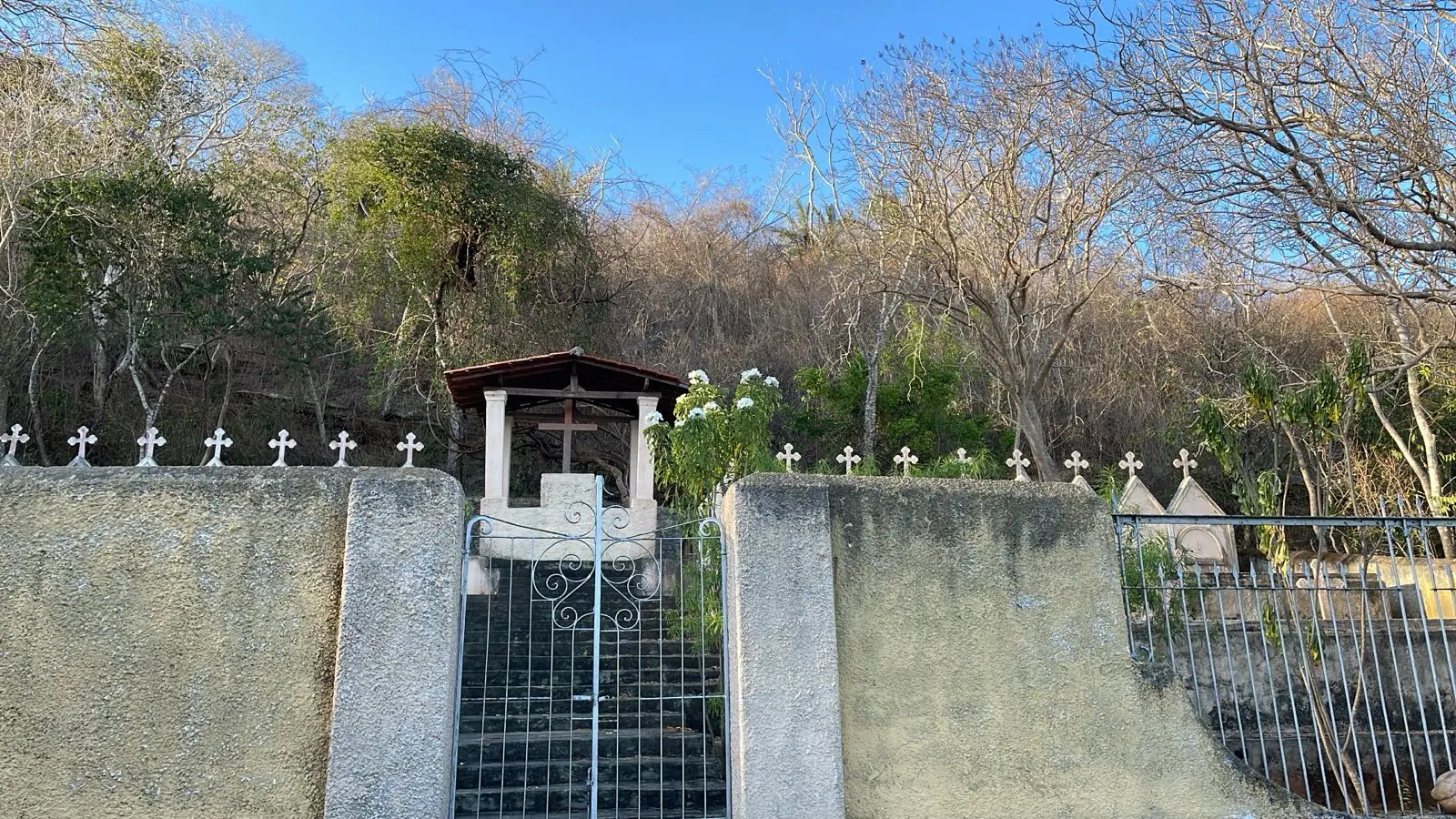 Cemitério com muro de cimento, grade de ferro e cruzes brancas em um ambiente ao ar livre, cercado por árvores e vegetação, sob céu azul.