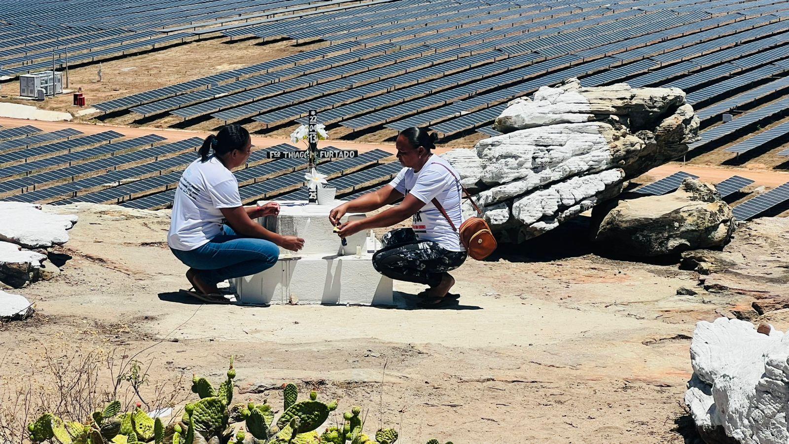 Duas mulheres ajoelhadas junto a um pequeno santuário no topo de um morro. Ao fundo, um vasto campo de painéis solares.