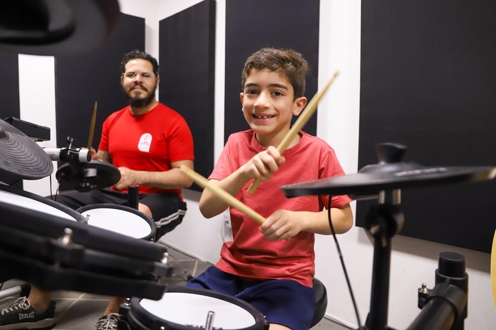 Na imagem, um professor e um aluno, ambos sentados e de blusa vermelha, compartilham ensinamentos sobre o instrumento musical bateria na escola GLB Music.