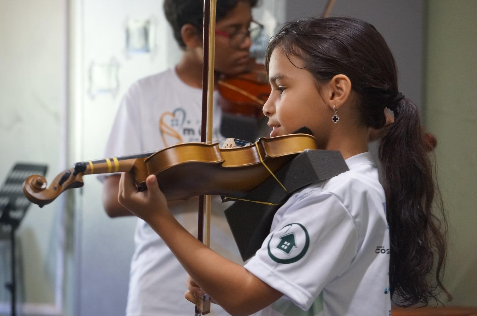 Na imagem, uma criança de blusa branca e cabelos amarrados aprende a tocar violino; ao fundo, outra criança, desta vez um menino, também aprende o instrumento na escola Casa de Vovó Dedé.