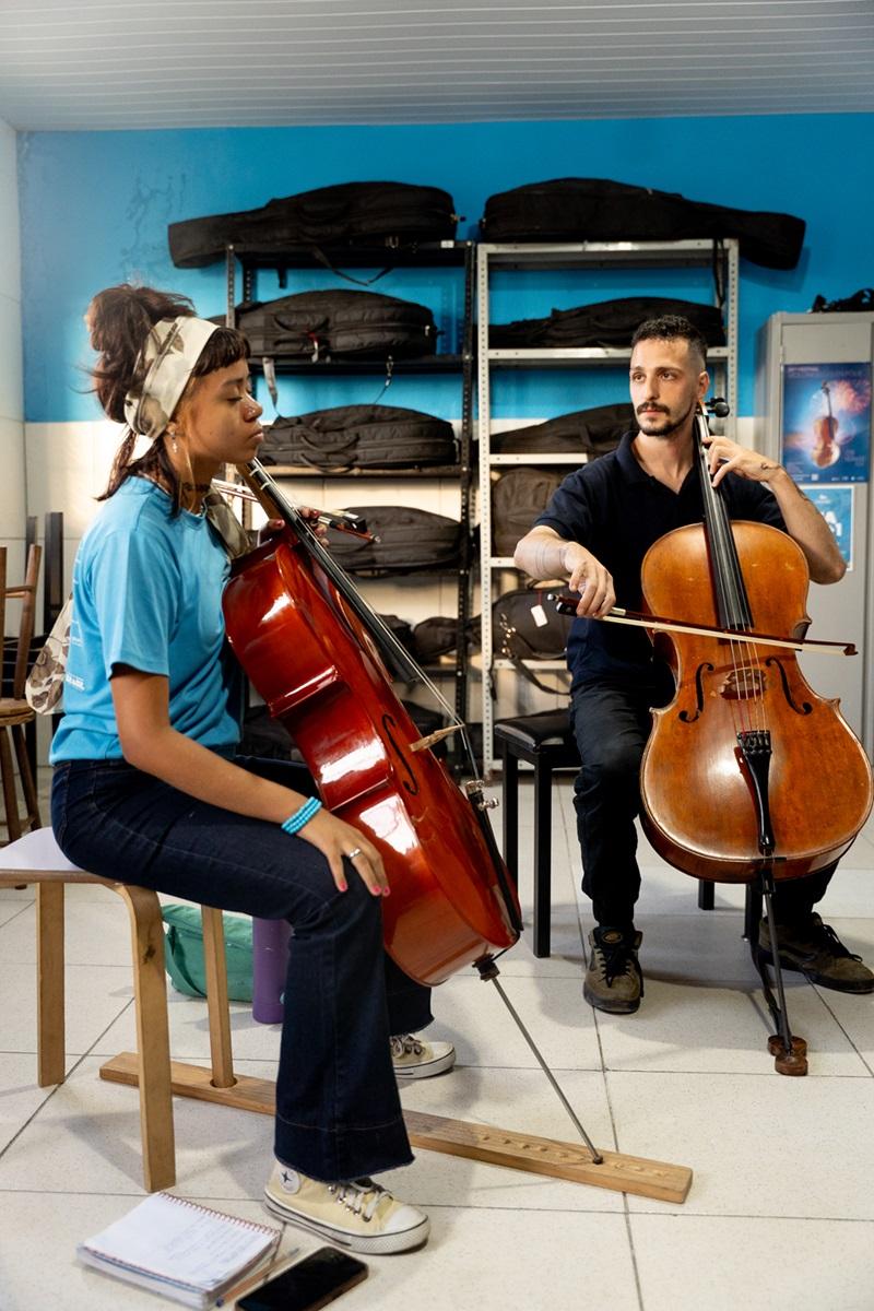 Na imagem, duas pessoas sentadas participam de aula de violoncelo; à esquerda, uma jovem negra, com blusa azul e cabelo amarrado; à direita, um homem branco, com camisa preta, ambos na escola Instituto Jacques Klein.