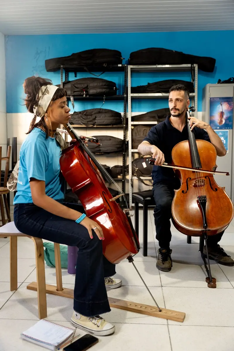 Na imagem, duas pessoas sentadas participam de aula de violoncelo; à esquerda, uma jovem negra, com blusa azul e cabelo amarrado; à direita, um homem branco, com camisa preta, ambos na escola Instituto Jacques Klein.
