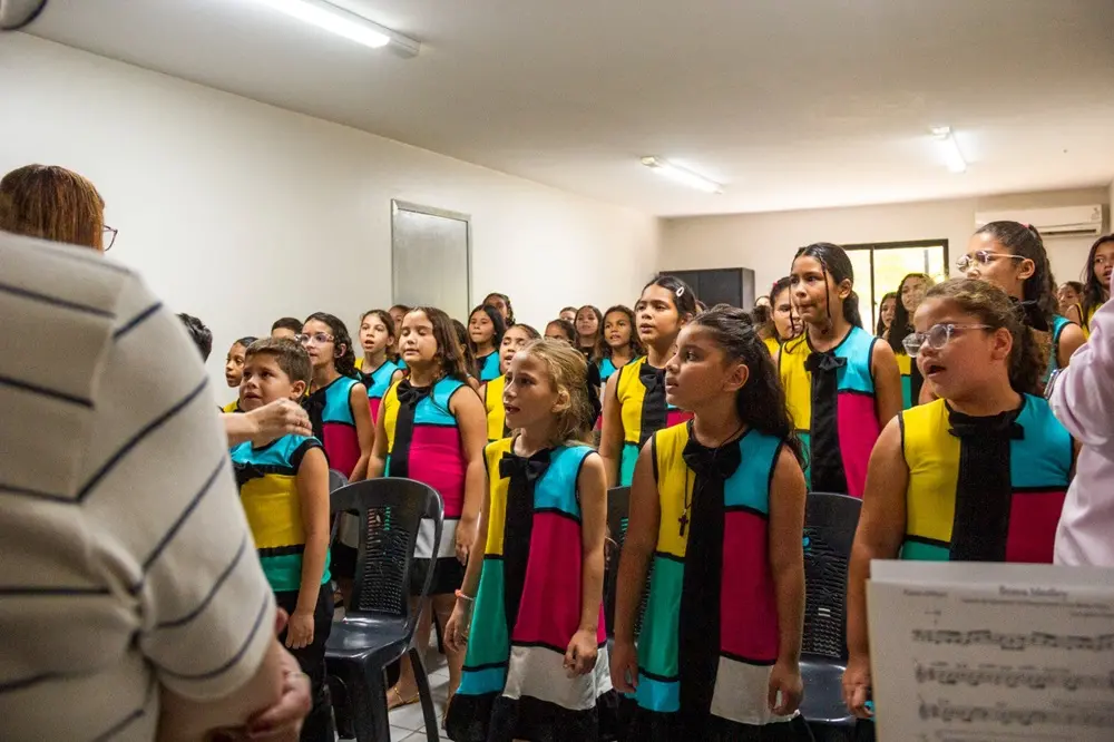 Na imagem, dezenas de crianças com vestes coloridas, em vermelho, verde, azul e amarelo, se apresentam no coral da escola Tapera das Artes.