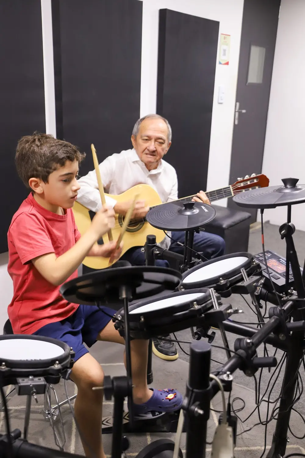 Na imagem, dois estudantes de música participam de uma aula; à esquerda, um menino de blusa vermelha toca bateria e, à direita, um idoso de blusa branca toca violão, ambos na escola GLB Music.