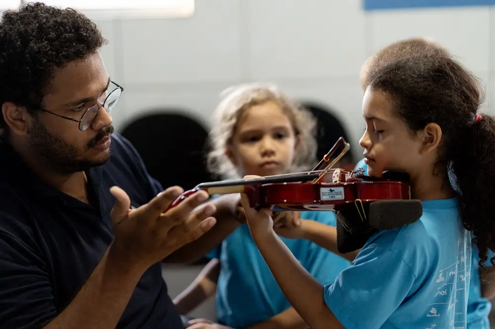 Na imagem, um professor e uma estudante dividem ensinamentos sobre violino durante aula de música no Instituto Jacques Klein; à esquerda, o professor tem pele negra, usa óculos e está de camisa preta; à direita, a estudante tem pele negra, está de cabelos amarrados e está com blusa azul.