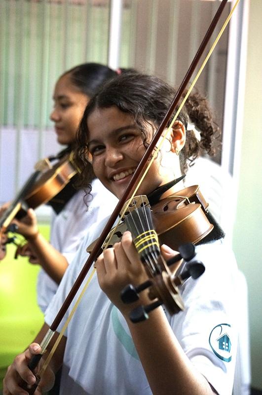 Na imagem, uma menina de pele negra, cabelos amarrados e sorrindo, toca violino durante aula na escola Casa de Vovó Dedé.