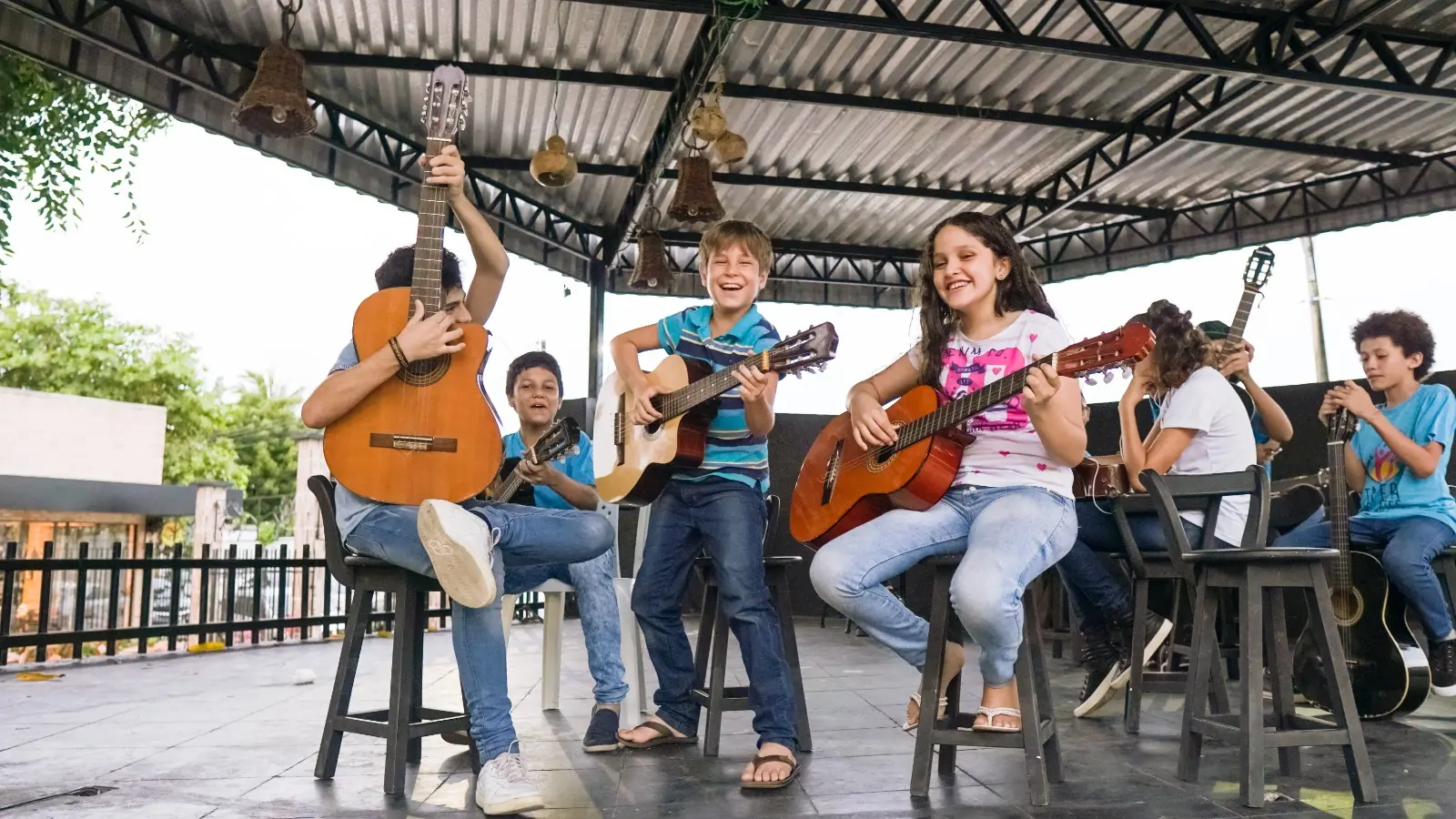 Na imagem, três crianças tocam diferentes instrumentos em amplo espaço na escola Tapera das Artes.