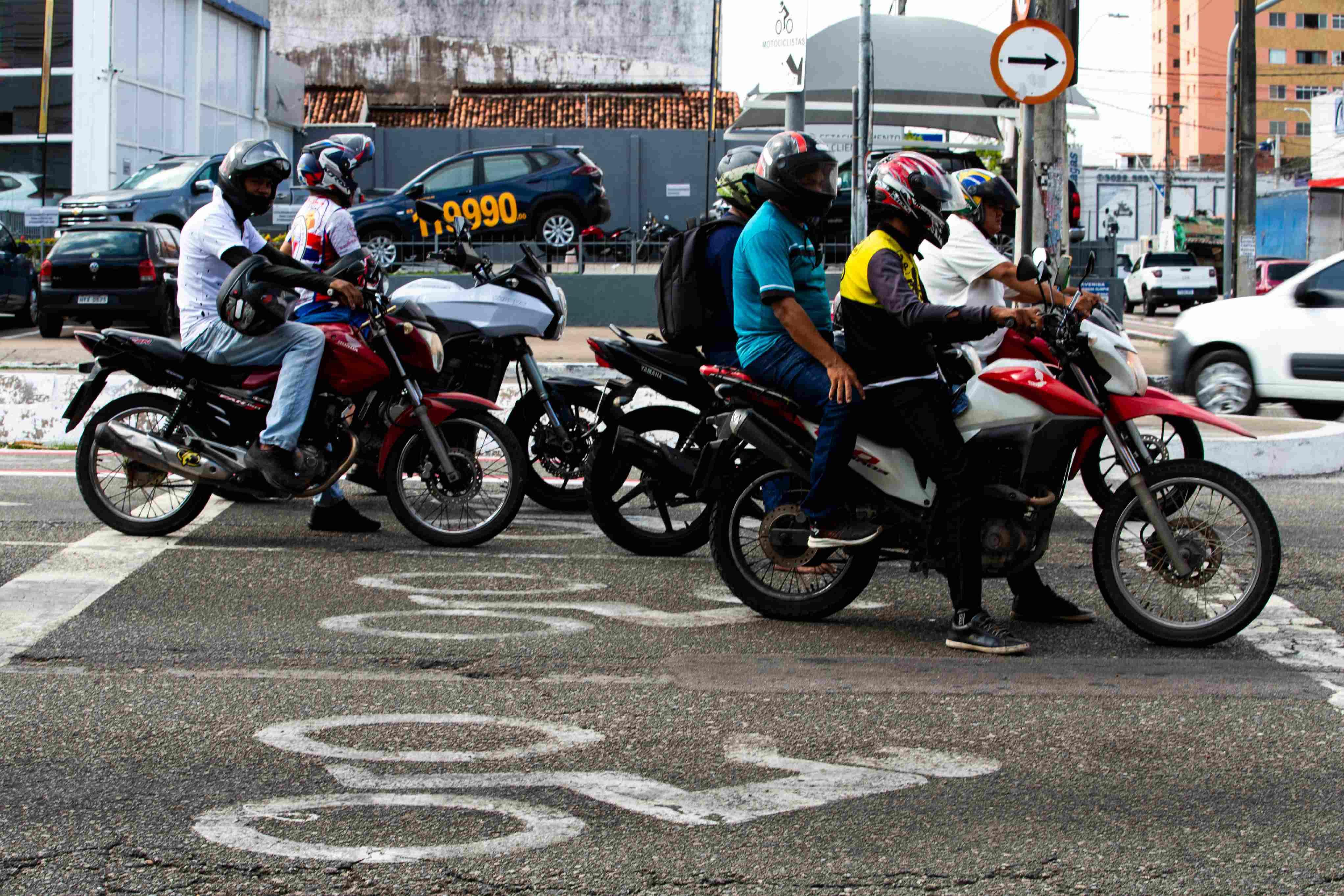 Grupo de motociclistas de capacete parados em uma faixa de retenção em um semáforo na cidade. Símbolo de bicicleta pintado no asfalto em primeiro plano.