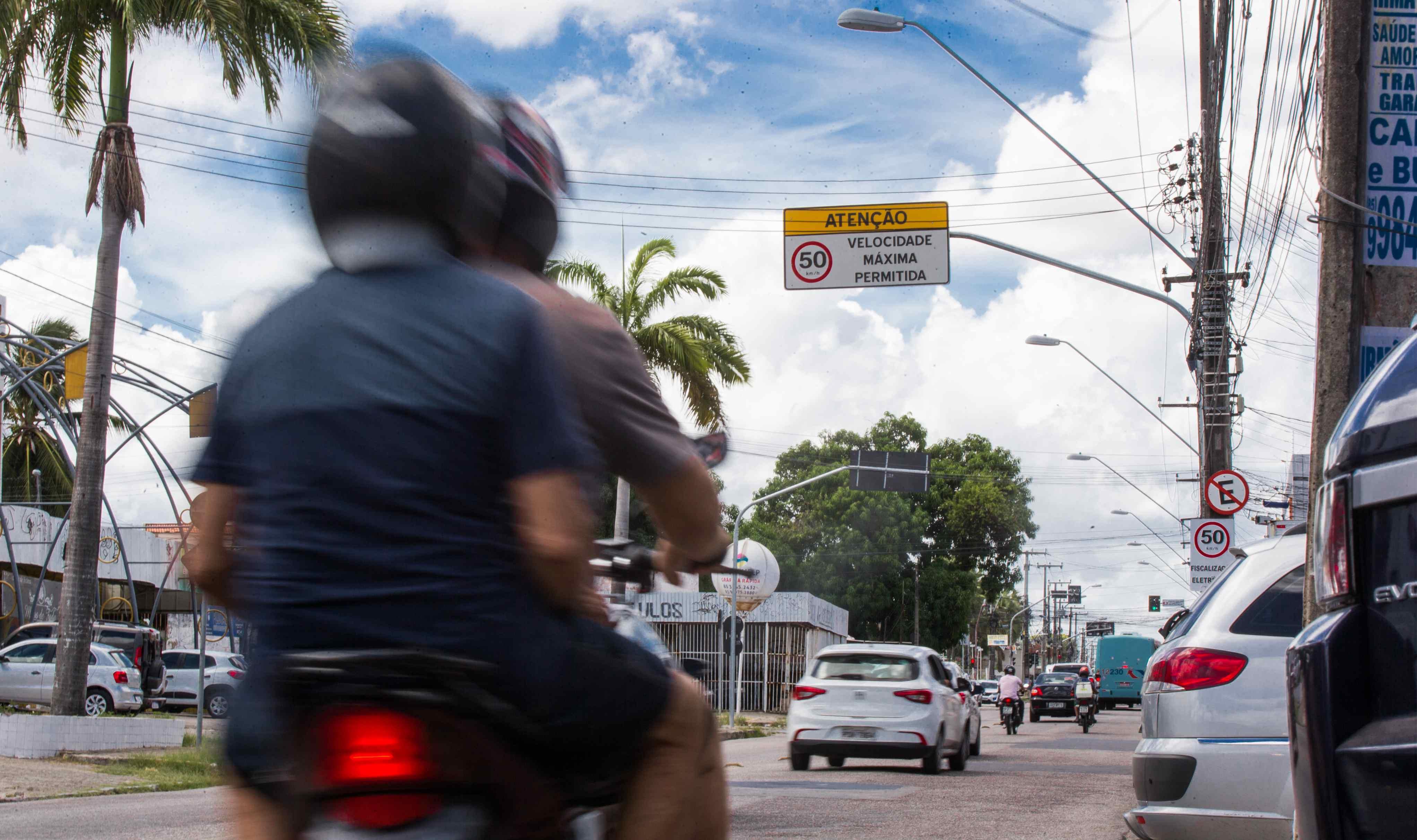 Motociclista borrado em primeiro plano em uma rua urbana. O foco está em um sinal de trânsito elevado no centro que indica “ATENÇÃO VELOCIDADE MÁXIMA PERMITIDA: 50 km/h”.