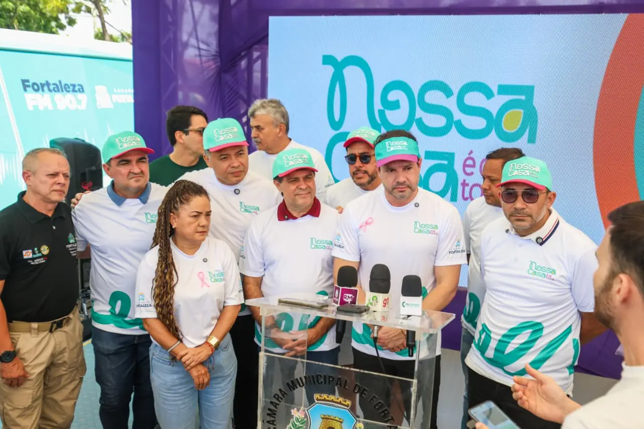 Grupo de pessoas participa de coletiva de imprensa durante evento do programa “Nossa Casa é Fortaleza”, usando camisetas e bonés com o nome da iniciativa, diante de painel colorido e púlpito com o brasão da Câmara Municipal de Fortaleza.