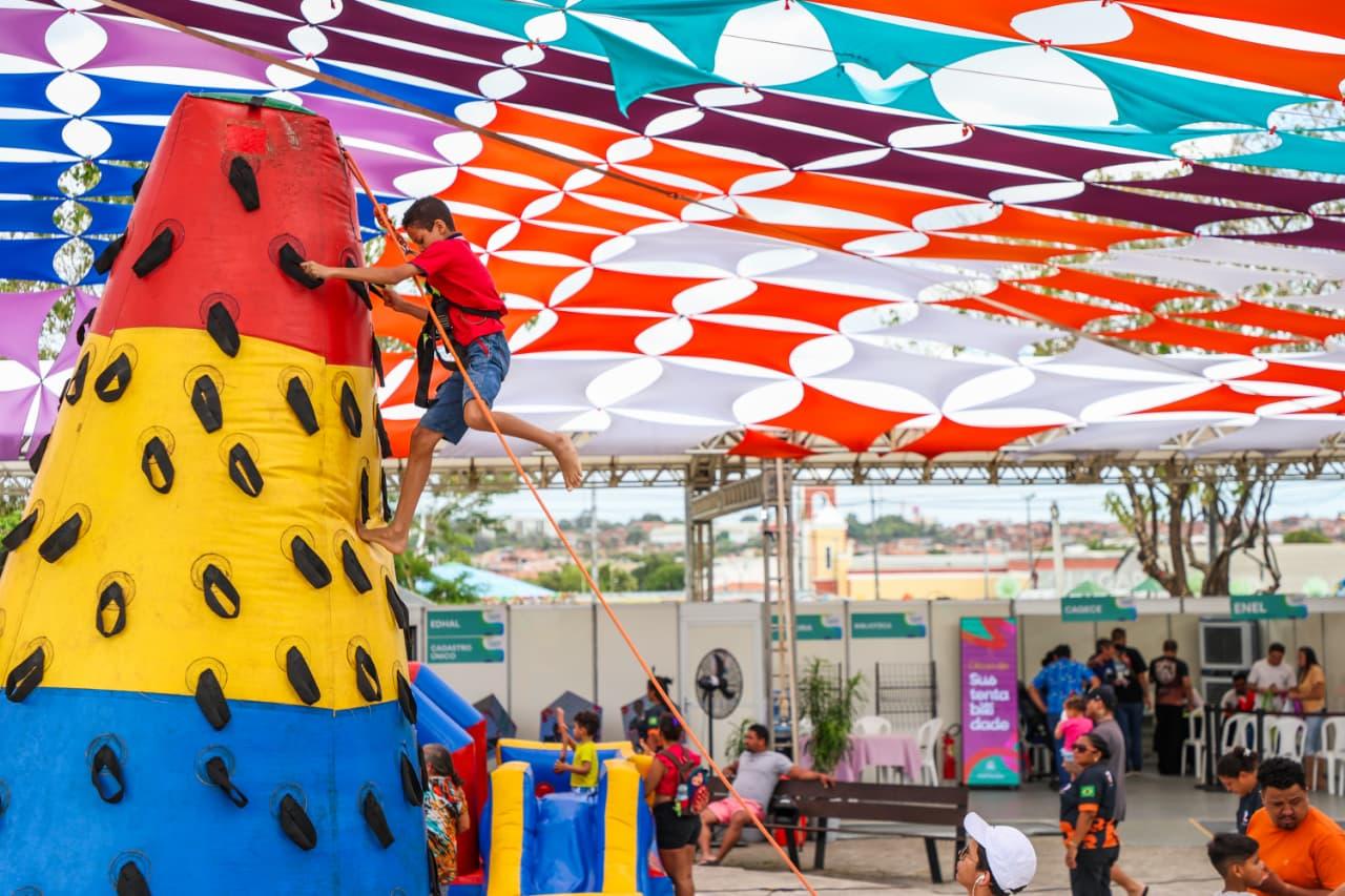 Criança escala estrutura inflável colorida em formato de cone, presa por corda de segurança, durante evento ao ar livre com tendas e cobertura de tecidos coloridos.