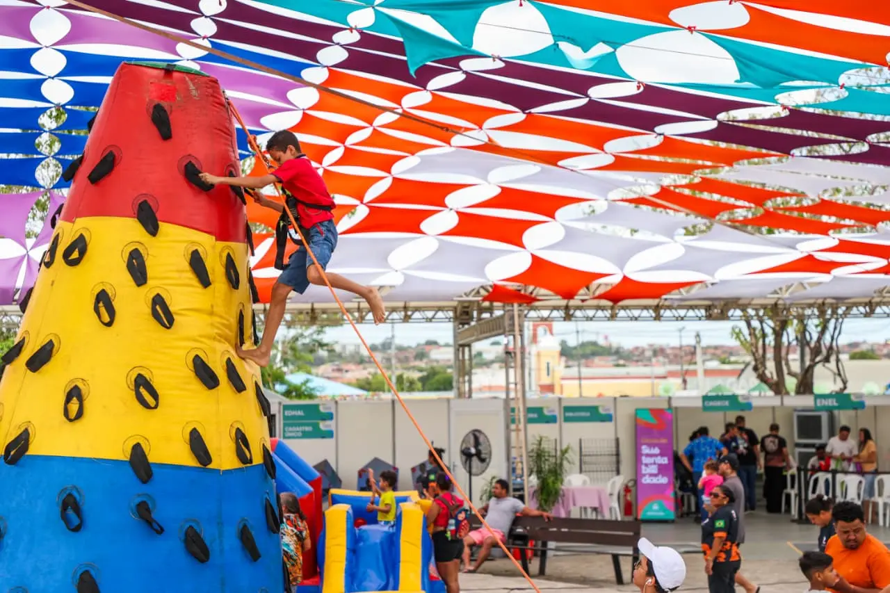 Criança escala estrutura inflável colorida em formato de cone, presa por corda de segurança, durante evento ao ar livre com tendas e cobertura de tecidos coloridos.
