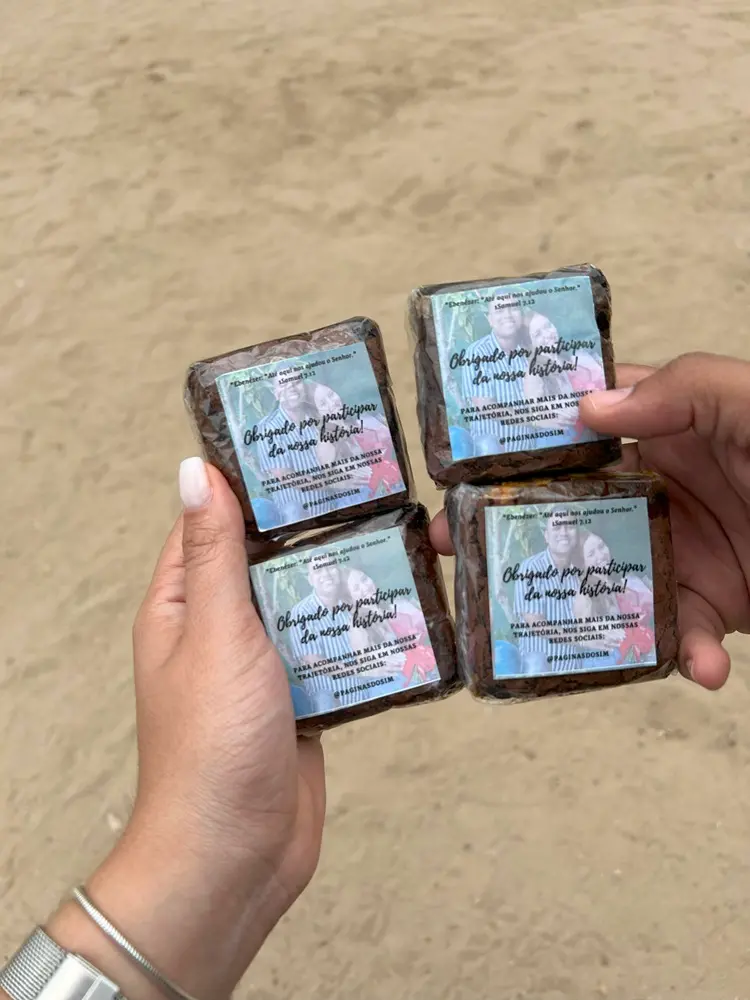 Na imagem, duas mãos seguram quatro brownies embalados individualmente. Os brownies estão dispostos em um quadrado dois por dois. Cada embalagem tem um adesivo personalizado com a foto de um casal sorrindo, um homem e uma mulher, em cima. O texto em português no adesivo diz: 