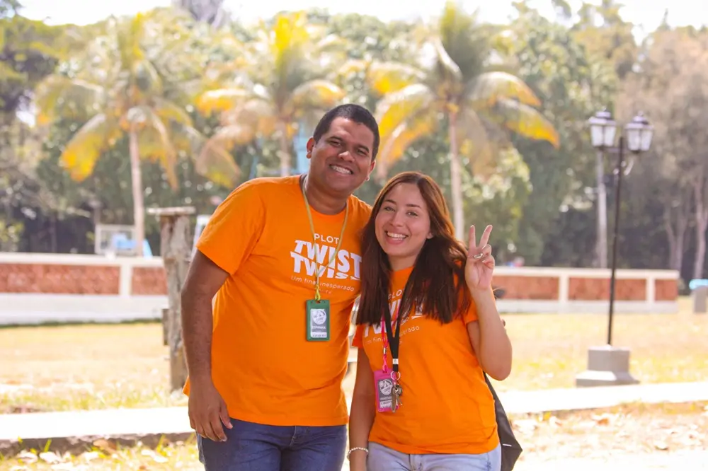 Na imagem, uma foto externa em um dia ensolarado mostra um casal, Ezequiel e Larissa, ambos vestindo camisetas laranja brilhante. Eles estão sorrindo para a câmera. O homem, Ezequiel, está à esquerda e tem pele morena escura e cabelo escuro curto, vestindo jeans escuro. A mulher, Larissa, está à direita e tem cabelos longos e castanhos, e está fazendo o sinal de 