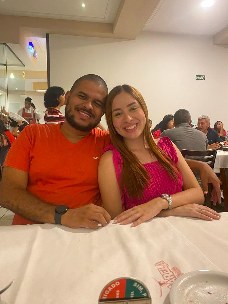 Na imagem, uma foto de casal tirada em um restaurante. Um homem de pele parda e barba, vestindo uma camisa polo laranja, está sentado à esquerda. Ele abraça uma mulher de cabelos ruivos e longos, vestida com um top rosa choque, sentada à direita. Ambos sorriem para a câmera. Eles estão sentados a uma mesa coberta com uma toalha branca. Ao fundo, outras pessoas jantam.