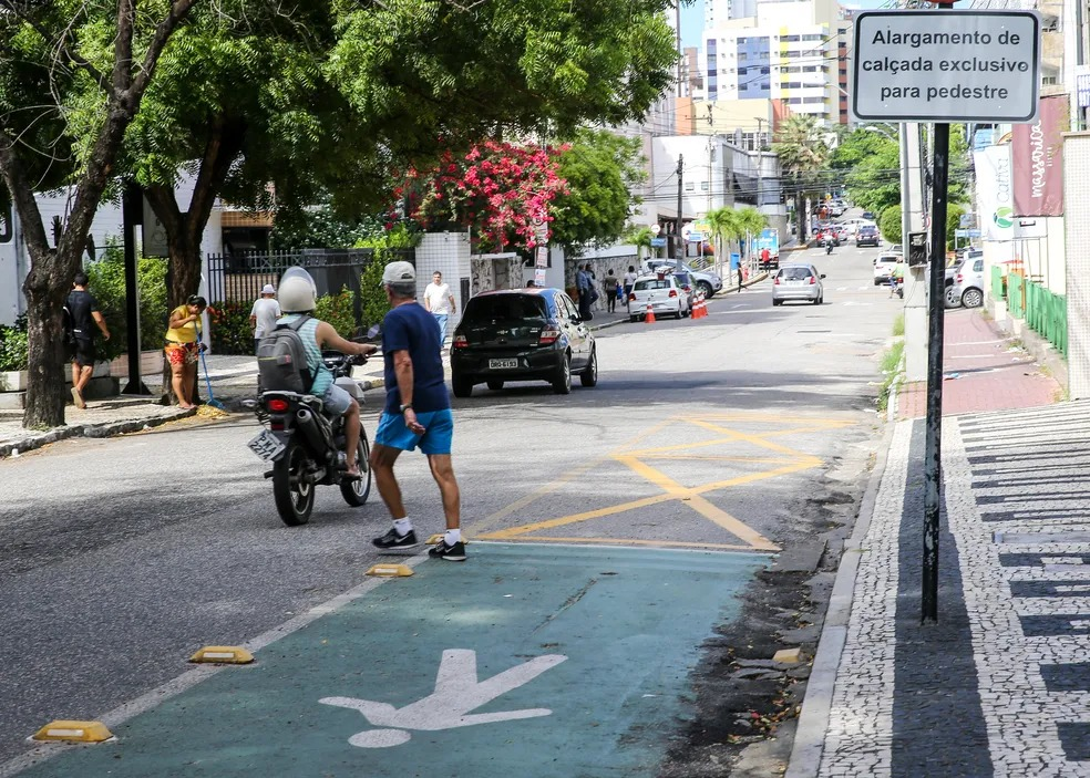 Cena de rua mostrando um “alargamento de calçada exclusivo para pedestre” pintado de verde-claro com sinalização. Motociclista e pedestre em primeiro plano, com trânsito e árvores ao fundo.
