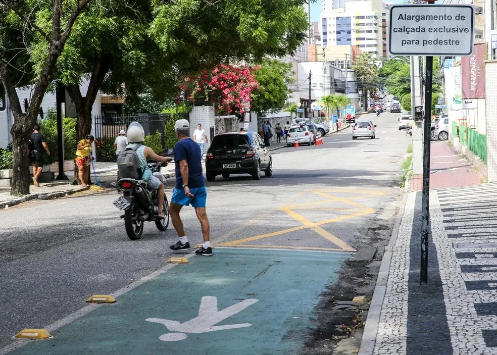 Cena de rua mostrando um “alargamento de calçada exclusivo para pedestre” pintado de verde-claro com sinalização. Motociclista e pedestre em primeiro plano, com trânsito e árvores ao fundo.