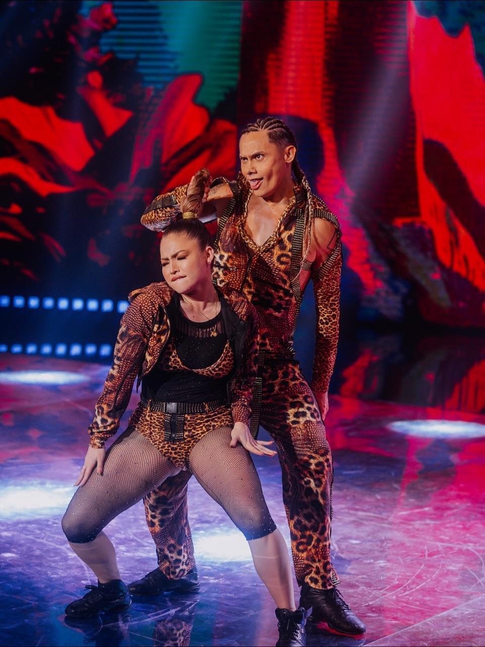 Na imagem, uma performance de dança. Duas pessoas, um homem e uma mulher, estão vestindo macacões e roupas combinando com estampa de onça (animal print) e detalhes brilhantes ou de strass. O homem, em pé, tem cabelo em tranças longas e uma expressão facial intensa, com a língua ligeiramente para fora. Ele está segurando o topo da cabeça com uma mão. A mulher, agachada em uma pose de dança, com os joelhos dobrados e os braços levemente afastados do corpo, está usando meias-calças arrastão (fishnet tights) e botas de cano curto. Ambos têm pele bronzeada e parecem estar no meio de uma rotina coreografada. Eles estão em um palco com piso brilhante, refletindo as luzes azuis e vermelhas de um fundo dramático e abstrato. O nome do arquivo sugere que o homem é Silvero Pereira e a foto é da 