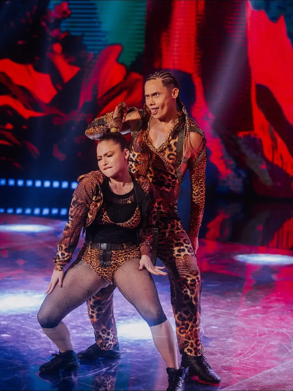 Na imagem, uma performance de dança. Duas pessoas, um homem e uma mulher, estão vestindo macacões e roupas combinando com estampa de onça (animal print) e detalhes brilhantes ou de strass. O homem, em pé, tem cabelo em tranças longas e uma expressão facial intensa, com a língua ligeiramente para fora. Ele está segurando o topo da cabeça com uma mão. A mulher, agachada em uma pose de dança, com os joelhos dobrados e os braços levemente afastados do corpo, está usando meias-calças arrastão (fishnet tights) e botas de cano curto. Ambos têm pele bronzeada e parecem estar no meio de uma rotina coreografada. Eles estão em um palco com piso brilhante, refletindo as luzes azuis e vermelhas de um fundo dramático e abstrato. O nome do arquivo sugere que o homem é Silvero Pereira e a foto é da 