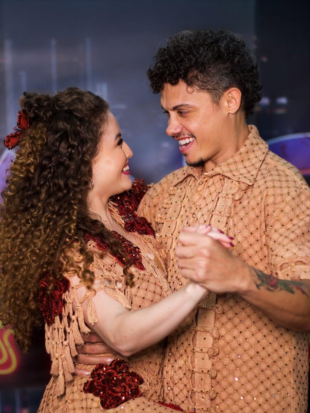 Na imagem, um casal de dançarinos. O homem, à direita, tem cabelo encaracolado escuro e está vestindo uma camisa de botão em um tom pêssego/marrom-claro, com textura que parece ser crochê ou tricô e pequenos brilhos. Ele tem tatuagens visíveis no braço esquerdo. A mulher, à esquerda, tem cabelo encaracolado castanho volumoso, decorado com um laço ou adorno vermelho. Ela está vestindo uma roupa que combina em cor com a camisa do homem, com franjas e apliques brilhantes em tom de vinho ou vermelho escuro. Os dois estão segurando as mãos e se olhando com sorrisos largos e olhares afetuosos, como se estivessem no meio de uma dança de salão. O fundo é ligeiramente desfocado, com tons escuros e alguns pontos coloridos, sugerindo um palco ou estúdio de dança. O nome do arquivo sugere que o homem é Silvero Pereira e a foto é da 