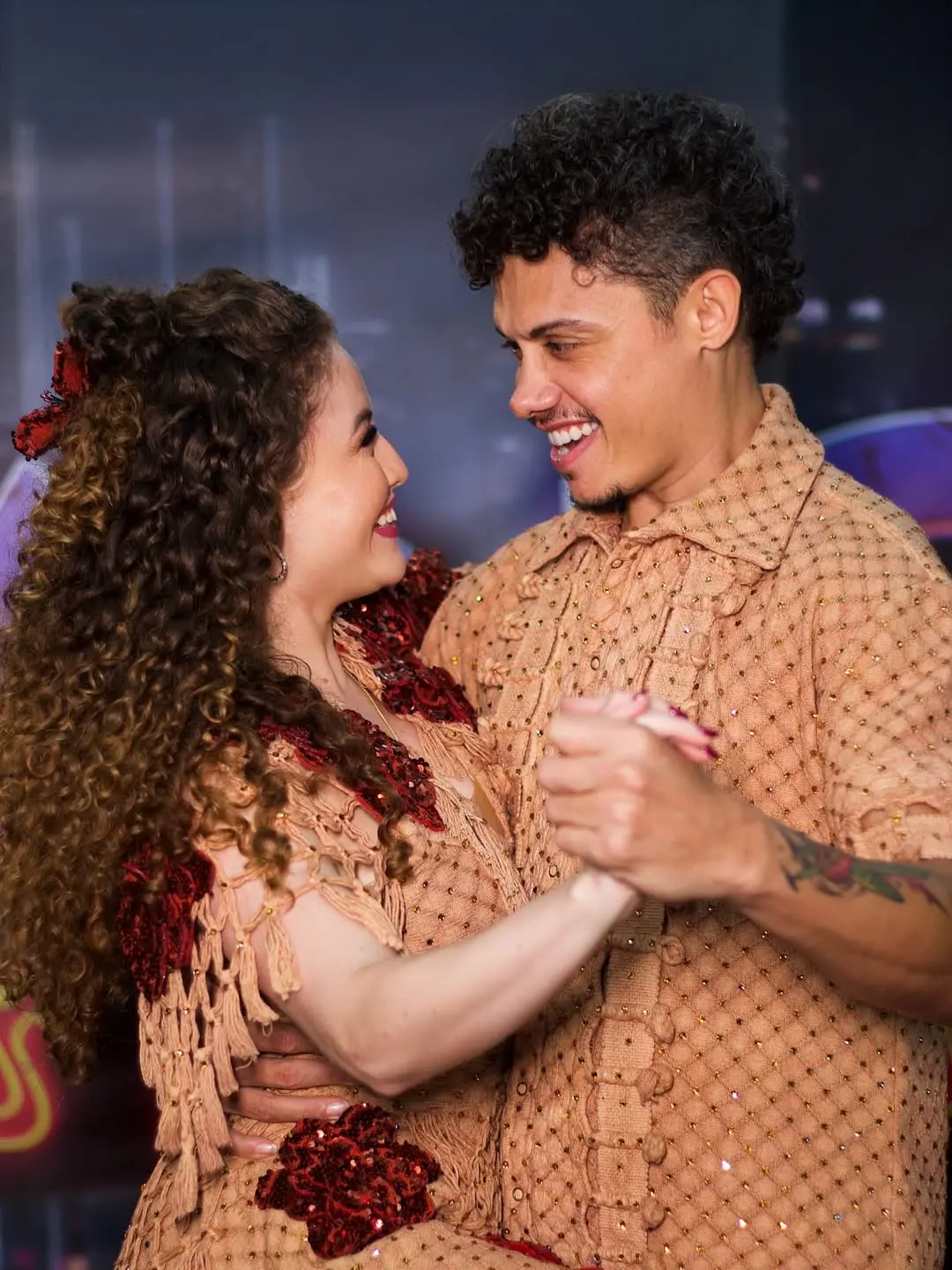 Na imagem, um casal de dançarinos. O homem, à direita, tem cabelo encaracolado escuro e está vestindo uma camisa de botão em um tom pêssego/marrom-claro, com textura que parece ser crochê ou tricô e pequenos brilhos. Ele tem tatuagens visíveis no braço esquerdo. A mulher, à esquerda, tem cabelo encaracolado castanho volumoso, decorado com um laço ou adorno vermelho. Ela está vestindo uma roupa que combina em cor com a camisa do homem, com franjas e apliques brilhantes em tom de vinho ou vermelho escuro. Os dois estão segurando as mãos e se olhando com sorrisos largos e olhares afetuosos, como se estivessem no meio de uma dança de salão. O fundo é ligeiramente desfocado, com tons escuros e alguns pontos coloridos, sugerindo um palco ou estúdio de dança. O nome do arquivo sugere que o homem é Silvero Pereira e a foto é da 