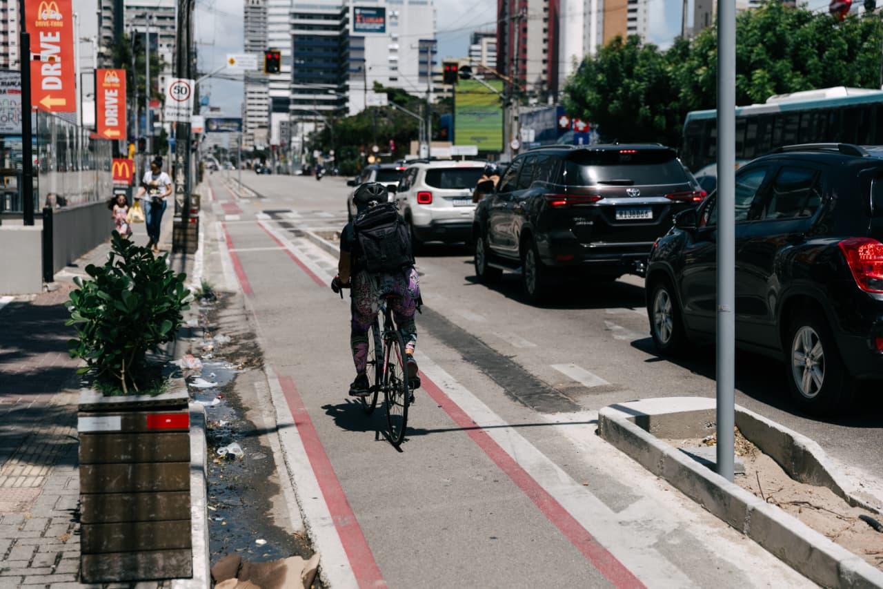 Ciclista de capacete e mochila pedalando em uma faixa segregada delimitada por linhas vermelhas. Trânsito de carros à direita e paisagem urbana com prédios altos ao fundo.