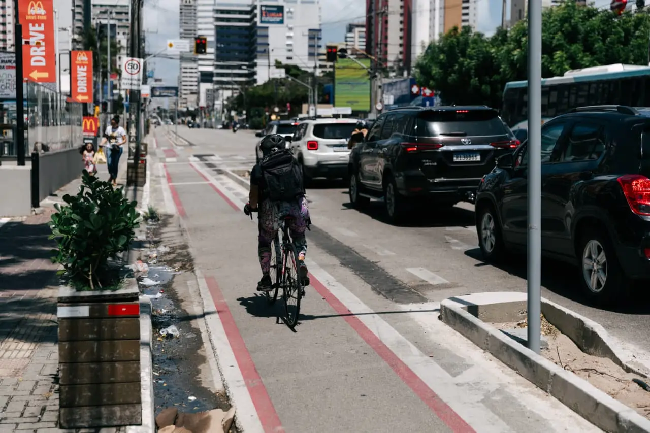 Ciclista de capacete e mochila pedalando em uma faixa segregada delimitada por linhas vermelhas. Trânsito de carros à direita e paisagem urbana com prédios altos ao fundo.