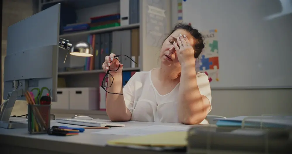 Na imagem, uma mulher sentada a uma mesa de escritório parece exausta ou estressada. Ela está vestindo uma blusa branca, tem a cabeça inclinada e está esfregando a testa e a pálpebra com a mão direita, enquanto segura um par de óculos na mão esquerda. A mesa de trabalho à sua frente tem um monitor de computador, um teclado, um mouse, uma luminária de mesa prateada e vários materiais de escritório, como um copo de lápis. O fundo mostra uma estante cheia de pastas e uma lousa branca. A iluminação é baixa e concentra-se na mesa e na mulher.