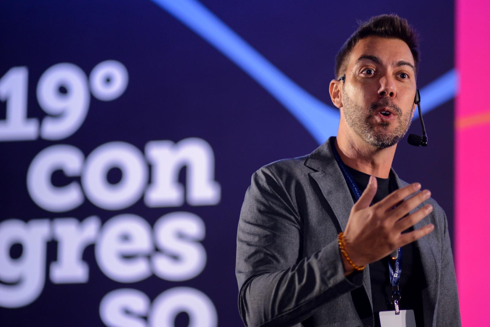 Na imagem, o psicólogo Lucas Freire, um homem de pele clara e barba, está em um palco escuro, falando com um microfone de cabeça. Ele está vestindo um paletó cinza sobre uma camiseta preta. Sua mão direita está levantada em um gesto de fala. O fundo tem um grande número 