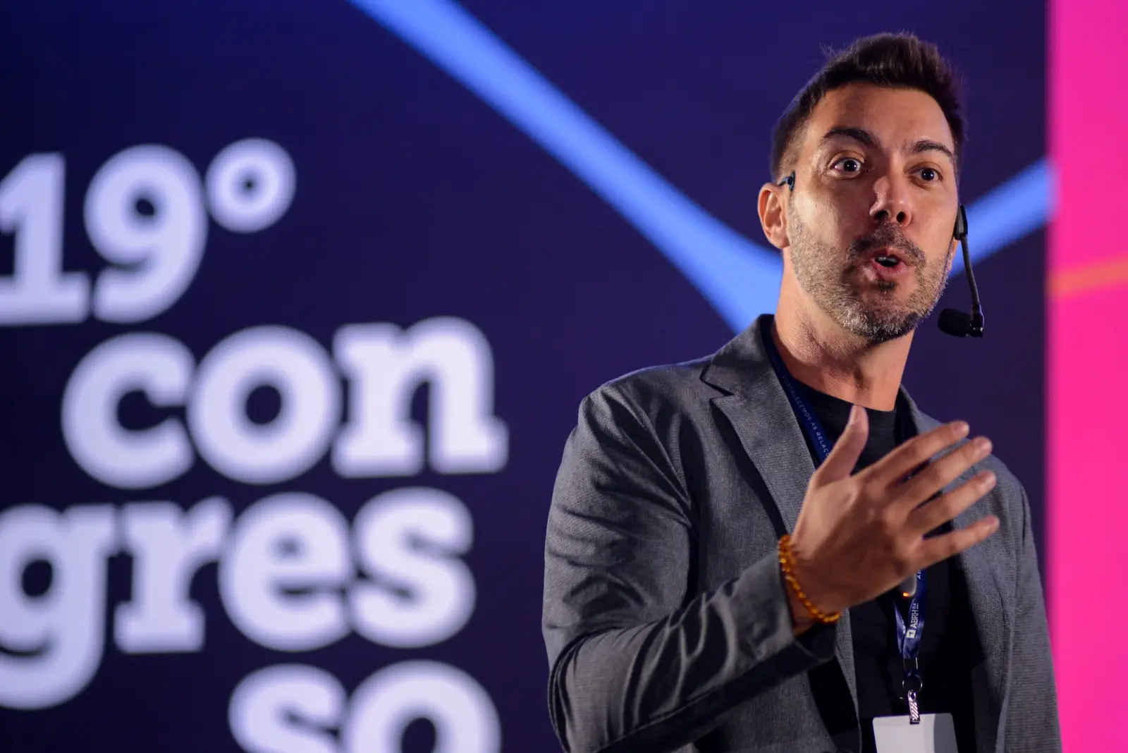 Na imagem, o psicólogo Lucas Freire, um homem de pele clara e barba, está em um palco escuro, falando com um microfone de cabeça. Ele está vestindo um paletó cinza sobre uma camiseta preta. Sua mão direita está levantada em um gesto de fala. O fundo tem um grande número 