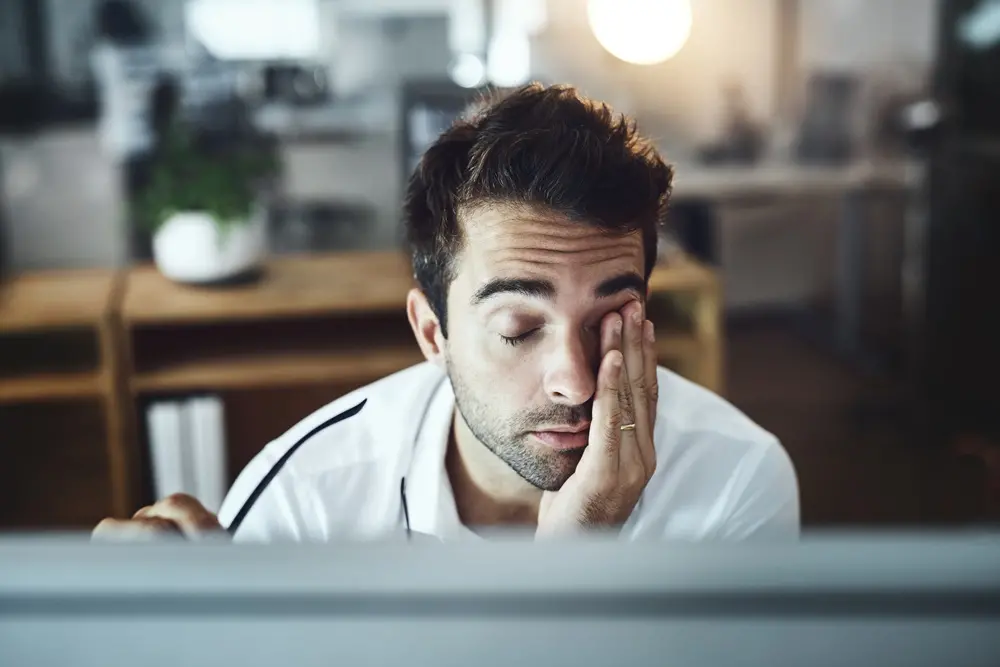 Na imagem, um homem com cabelo escuro e barba por fazer está sentado em uma mesa de escritório, parecendo exausto. Ele está vestindo uma camisa social branca e tem a cabeça apoiada na mão esquerda, cobrindo um dos olhos. Seus olhos estão fechados e ele parece estar descansando ou esfregando-os devido à fadiga. Um monitor de computador está em primeiro plano, parcialmente fora de foco. O fundo, também desfocado, mostra um escritório com prateleiras de madeira e uma planta. A iluminação é suave, com uma fonte de luz brilhante na parte superior central.
