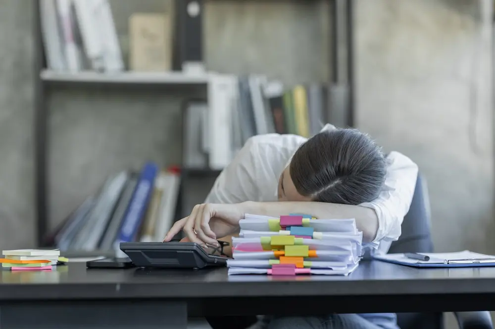 Na imagem, uma pessoa sentada em uma mesa de escritório está com a cabeça deitada sobre os braços cruzados, apoiados em uma pilha de documentos e pastas coloridas, o que sugere exaustão devido ao excesso de trabalho. Ela está vestindo uma camisa branca e sua mão direita está estendida, pressionando uma calculadora. A mesa é escura e também contém um smartphone, caneta e prancheta. Ao fundo, fora de foco, há uma estante de metal com livros e pastas.