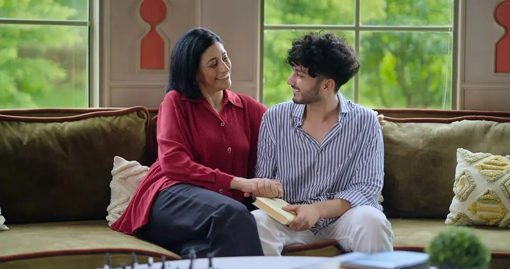 Na imagem, uma mulher mais velha de cabelos escuros e um jovem de cabelos cacheados e barba rala, ambos de etnia indiana, estão sentados em um sofá verde em uma sala com janelas grandes. A mulher, vestindo uma blusa vermelha e calças pretas, está sorrindo e olhando para o jovem. Ela tem a mão em cima da mão dele. O jovem está sorrindo para a mulher e segurando um livro com a outra mão. Eles parecem ser mãe e filho. O fundo mostra janelas com uma paisagem verde.