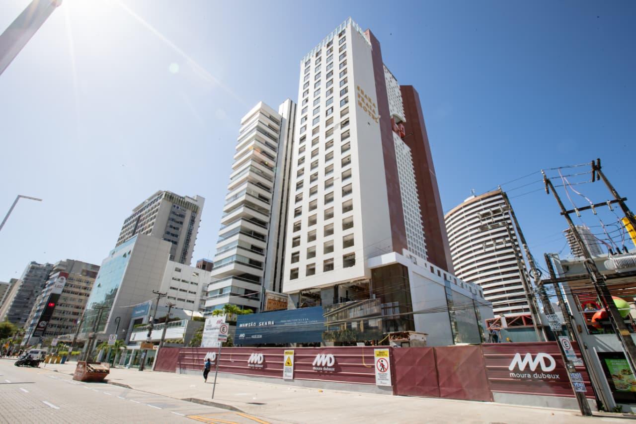 Vista ampla de um quarteirão com prédios altos, incluindo o Seara Praia Hotel em destaque, cercado por tapumes de obra da construtora Moura Dubeux, em uma avenida movimentada e céu ensolarado.