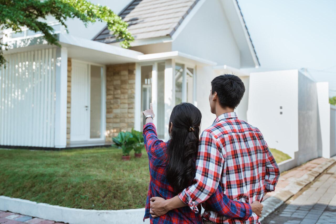 Casal de homem e mulher brancos, de cabelos pretos, de costas, olhando uma casa de arquitetura moderna e elegante, com fachada de alvenaria e grandes janelas.