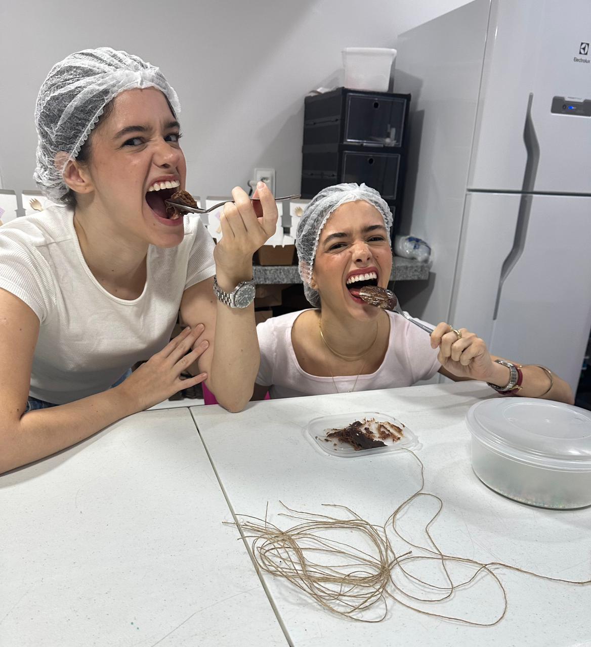 imagem mostra irmãs gêmeas que criaram empresa de bolo de chocolate, no ceará. elas estão com roupas claras, vestem toucas e colocam pedaços de bolo na boca