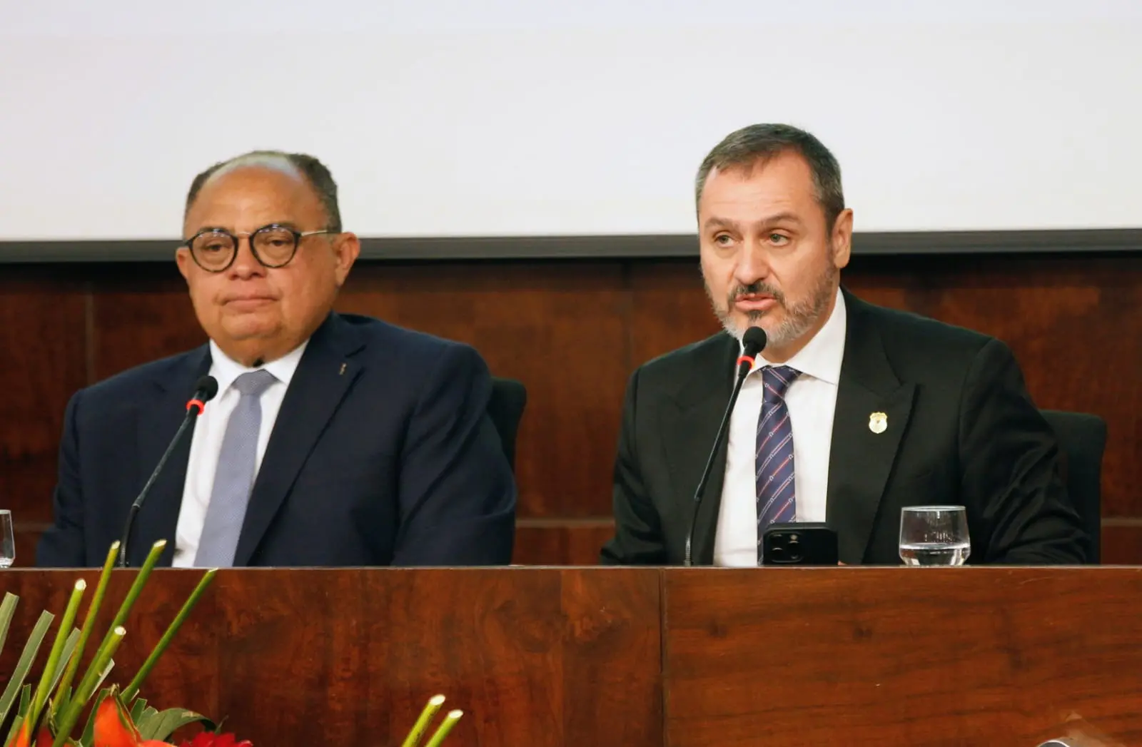Dois homens vestidos com terno e gravata participam de uma mesa de debates em um auditório. O homem à esquerda, de óculos, observa em silêncio; o à direita fala ao microfone, com expressão séria. À frente deles, sobre a mesa de madeira, há copos de água e arranjos de flores tropicais.