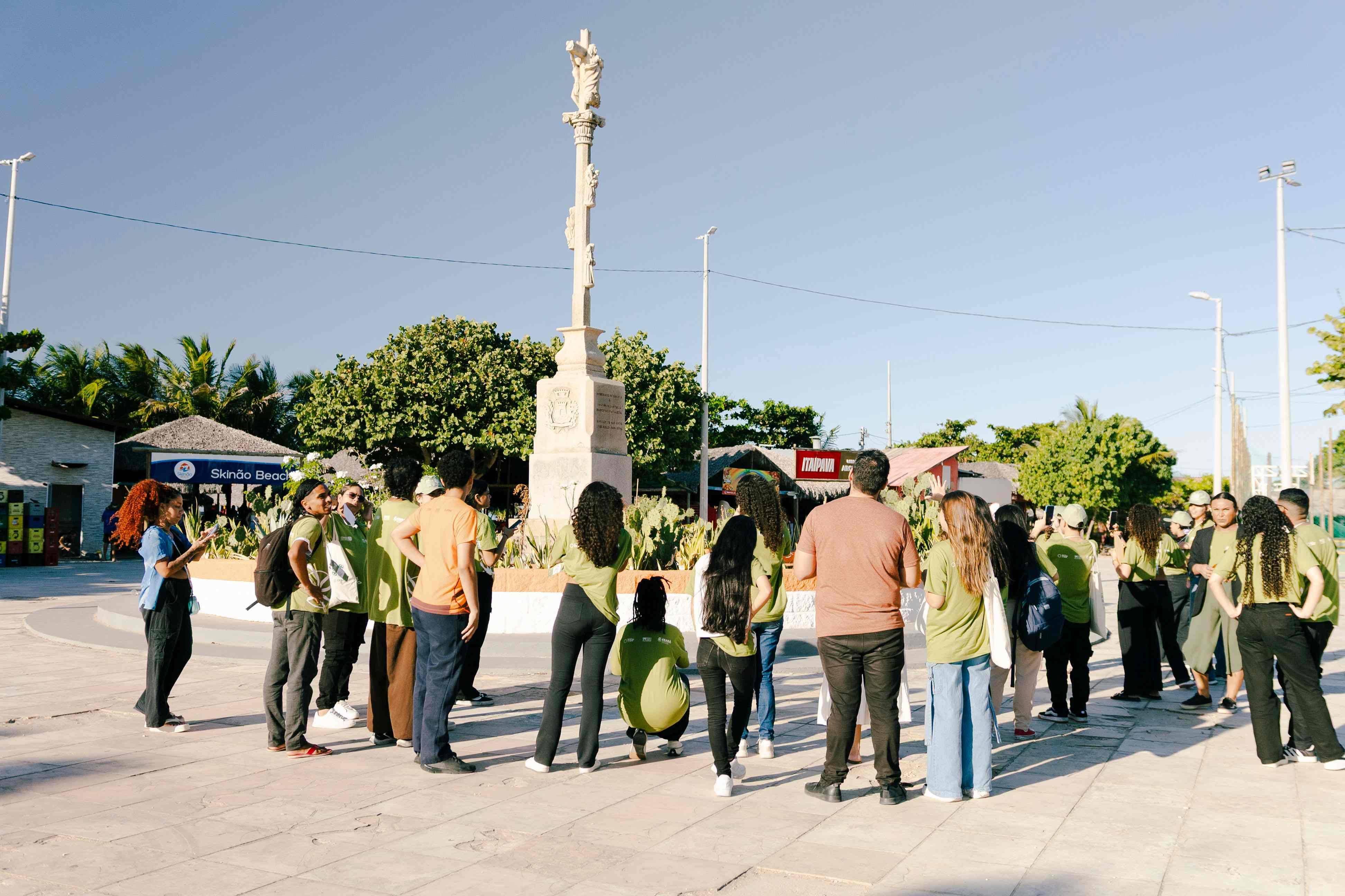 Grupo de pessoas reunido ao redor de um monumento de pedra em uma praça ao ar livre, com céu claro, árvores e quiosques ao fundo.