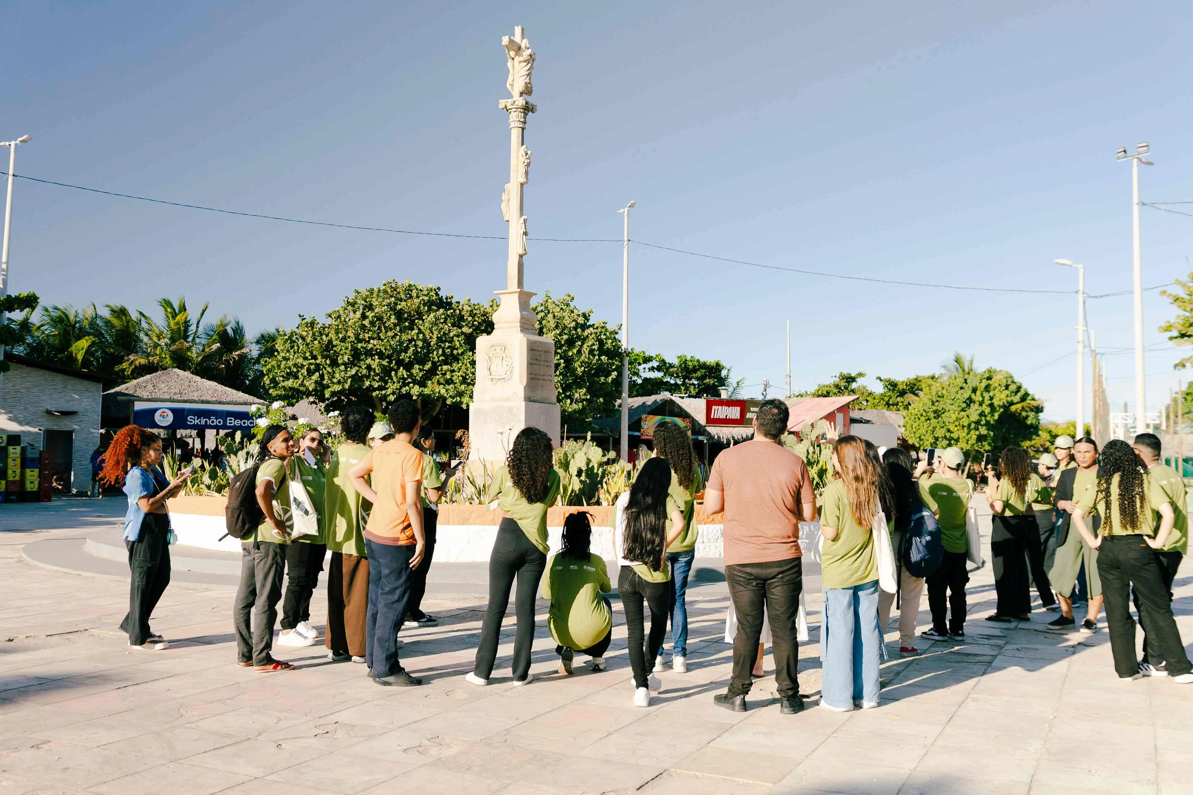 Grupo de pessoas reunido ao redor de um monumento de pedra em uma praça ao ar livre, com céu claro, árvores e quiosques ao fundo.