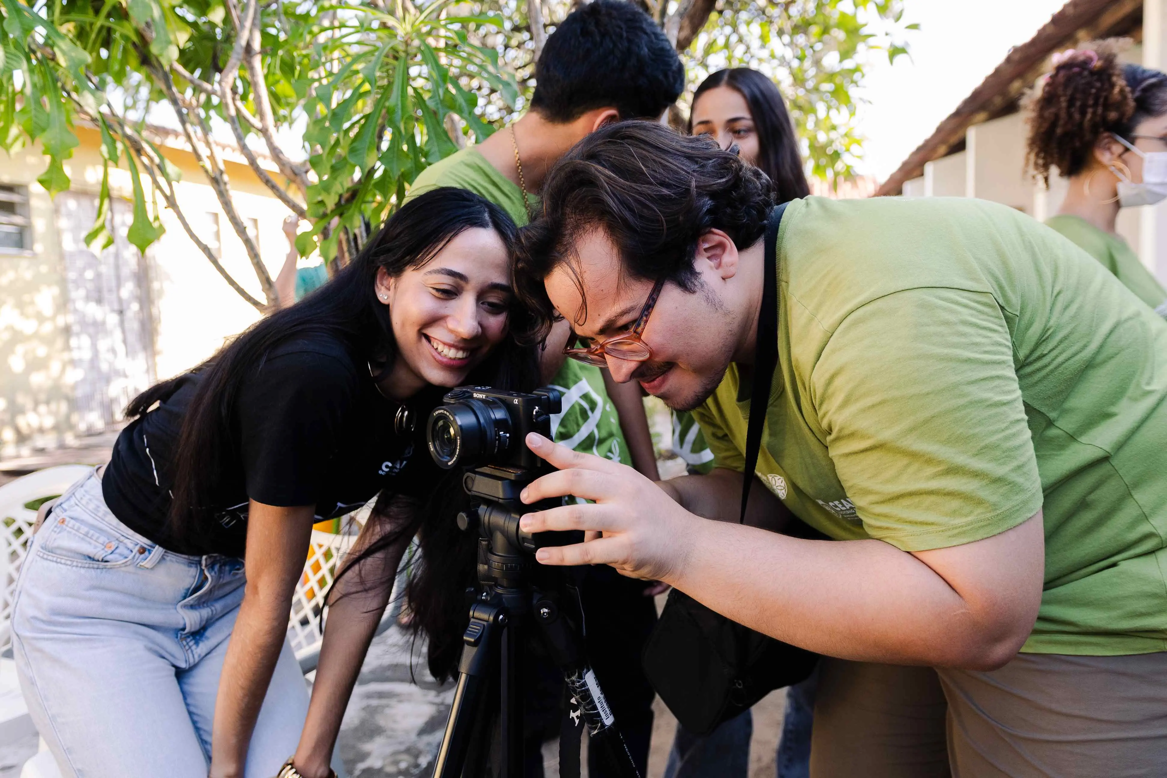 Jovens sorrindo enquanto observam uma câmera fotográfica em um tripé, em ambiente externo com árvores ao fundo.