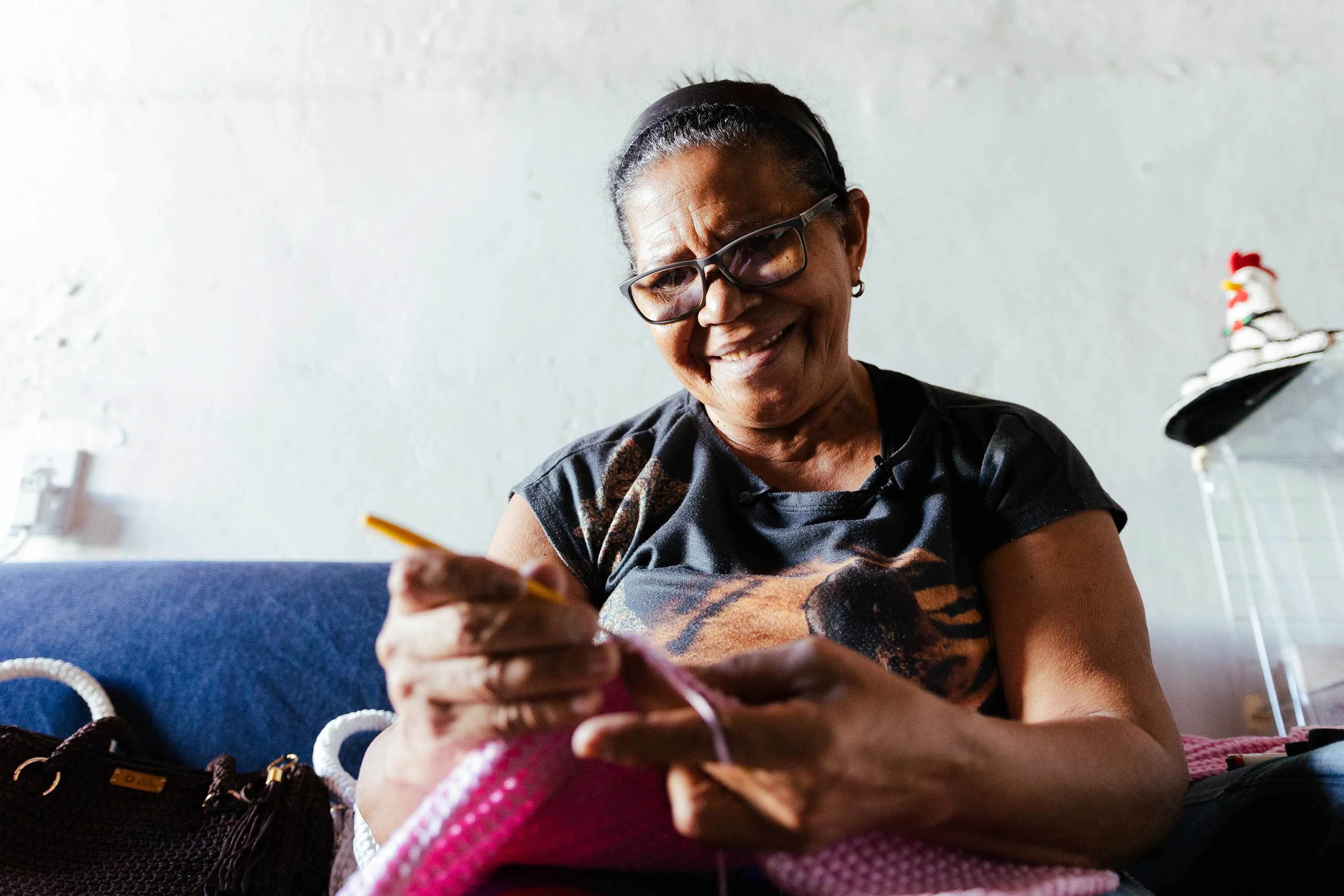 Close-up de Dona Francy, uma mulher sorridente de óculos, fazendo crochê com um tecido rosa vibrante. Ela está sentada em frente a uma parede branca e um sofá azul-escuro.