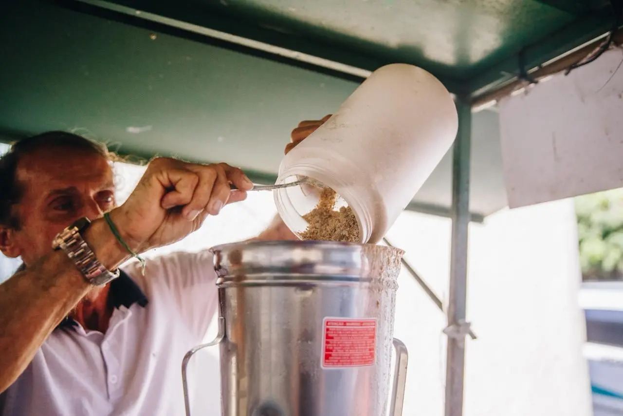 Na imagem, uma foto em close-up mostra um homem de camisa polo branca e cabelo escuro amarrado, adicionando um ingrediente em pó claro e granulado de um pote cilíndrico branco para o liquidificador industrial de metal prateado. Ele está segurando o pote com a mão esquerda e uma colher de metal com a direita para raspar o ingrediente. O pote está inclinado diretamente sobre a abertura do liquidificador. O topo de uma cobertura ou toldo verde escuro é visível no canto superior esquerdo. Um adesivo retangular vermelho com letras brancas no liquidificador diz 