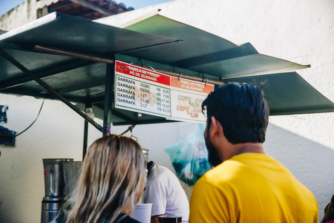 Na imagem, uma foto ligeiramente de baixo para cima e de trás mostra dois clientes olhando para a tabela de preços do 