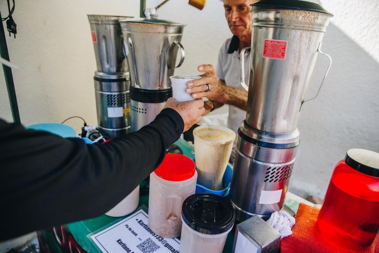 Na imagem, uma foto em close-up mostra uma transação no balcão de um quiosque de guaraná batido. Um cliente, usando uma blusa preta de manga comprida (apenas o braço e a mão são visíveis em primeiro plano, à esquerda), está estendendo a mão para receber um copo branco de guaraná batido. O homem atrás do balcão, que tem cabelos escuros e usa uma camisa polo branca, está entregando o copo. No balcão verde, há vários liquidificadores industriais de metal prateado (um com um adesivo vermelho de 