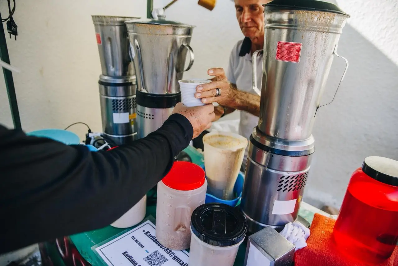 Na imagem, uma foto em close-up mostra uma transação no balcão de um quiosque de guaraná batido. Um cliente, usando uma blusa preta de manga comprida (apenas o braço e a mão são visíveis em primeiro plano, à esquerda), está estendendo a mão para receber um copo branco de guaraná batido. O homem atrás do balcão, que tem cabelos escuros e usa uma camisa polo branca, está entregando o copo. No balcão verde, há vários liquidificadores industriais de metal prateado (um com um adesivo vermelho de 