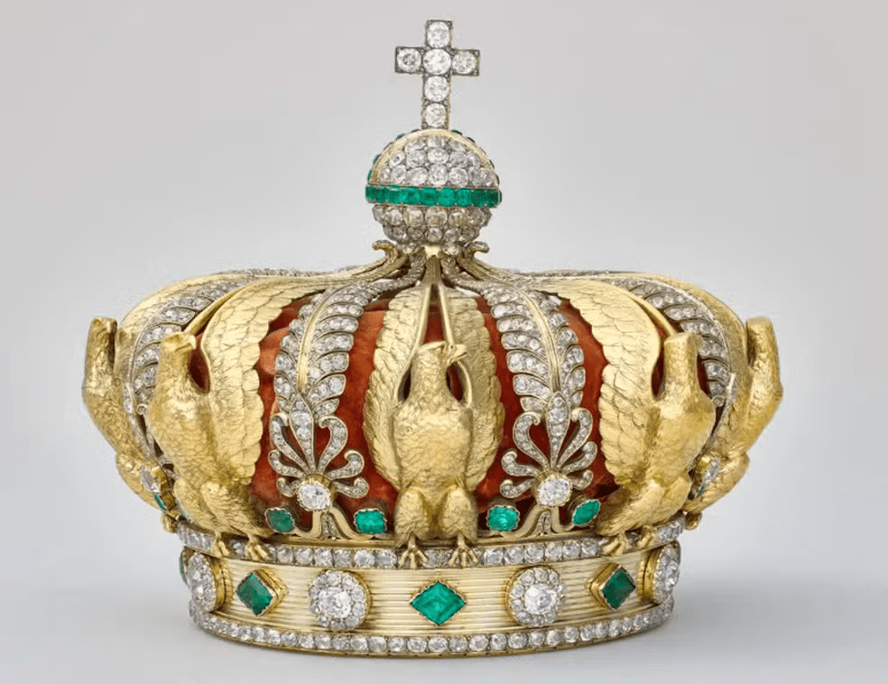 Coroa dourada ornamentada com diamantes e esmeraldas sobre um fundo neutro. A peça apresenta estrutura detalhada com águias em relevo e arabescos decorativos, além de uma faixa circular cravejada de pedras preciosas verdes e brancas. No topo, há um globo coberto de diamantes com uma cruz igualmente adornada. O interior da coroa é revestido por tecido vermelho aveludado, visível entre os arcos metálicos. Imagem usada em matéria sobre roubo no Museu do Louvre.