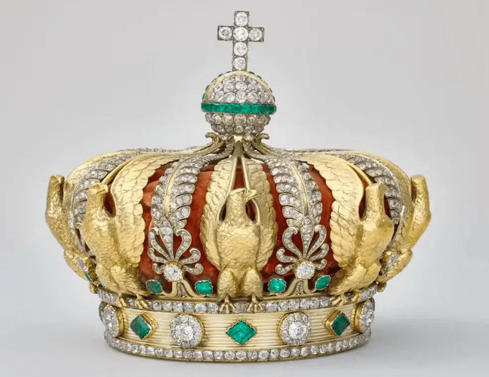 Coroa dourada ornamentada com diamantes e esmeraldas sobre um fundo neutro. A peça apresenta estrutura detalhada com águias em relevo e arabescos decorativos, além de uma faixa circular cravejada de pedras preciosas verdes e brancas. No topo, há um globo coberto de diamantes com uma cruz igualmente adornada. O interior da coroa é revestido por tecido vermelho aveludado, visível entre os arcos metálicos. Imagem usada em matéria sobre roubo no Museu do Louvre.